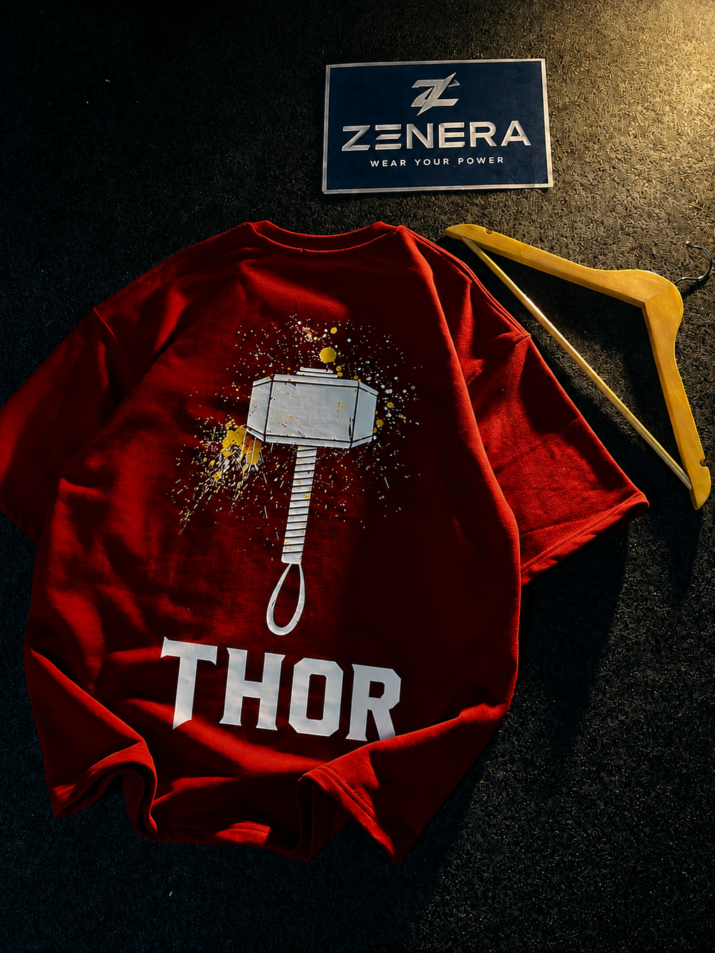 A red short-sleeve T-shirt laid flat on a dark textured surface, featuring a large hammer graphic with paint splatter and the word 'THOR' in bold white letters. A yellow hanger rests beside the shirt, and a ZENERA brand tag is visible above it.